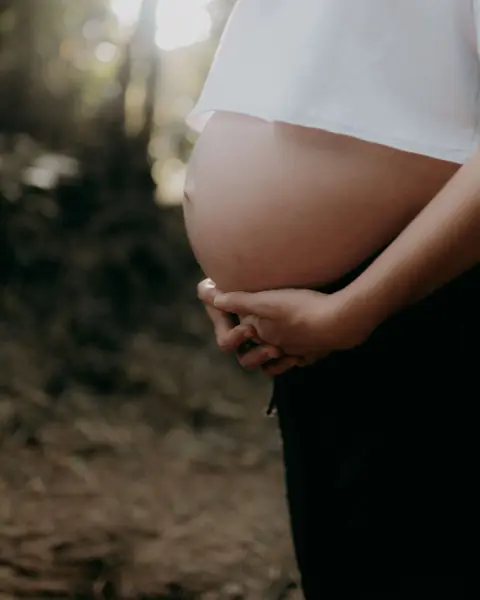 Close-up of a pregnant woman cradling her belly with both hands in soft forest light